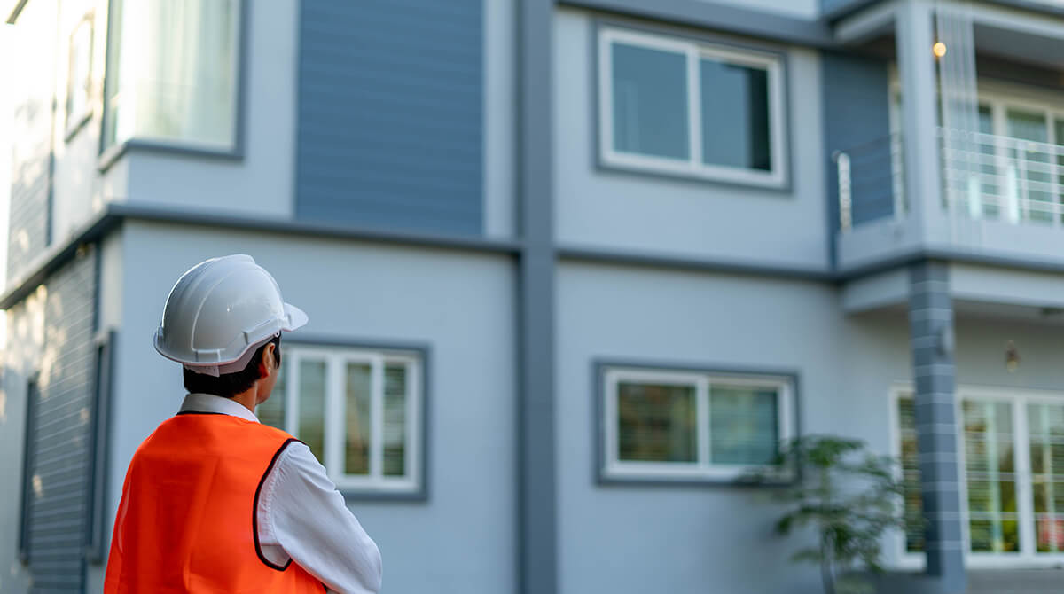 Construction worker wearing a white hard hat and orange safety vest inspecting a modern gray house exterior.