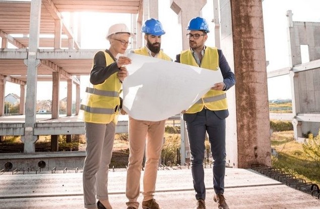Three construction professionals wearing safety helmets and reflective vests examining blueprints on a construction site.