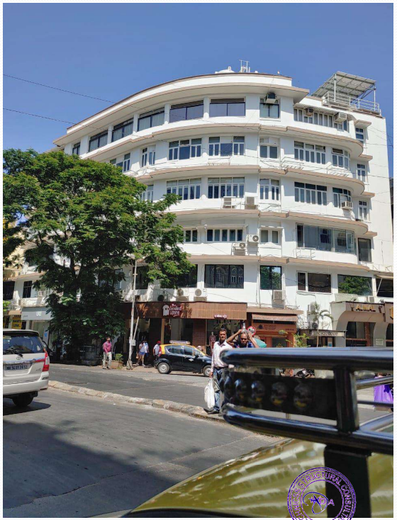 White multi-story building with large windows on a sunny day, with people walking and vehicles on the street in front.