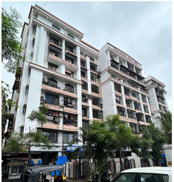 Multi-story white residential building with balconies, trees in front, and vehicles parked at street level under cloudy sky.