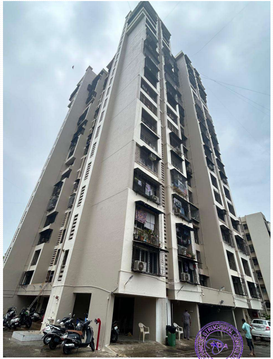 Tall residential building with multiple balconies under a cloudy sky and parked scooters and cars at its base.