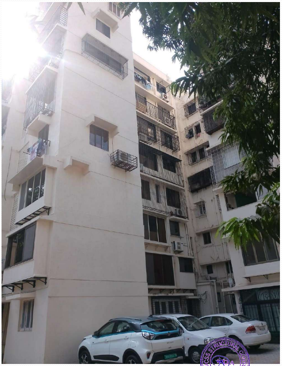 Multi-story residential building with multiple windows and balconies, parked cars in front, and sunlight filtering through tree branches.
