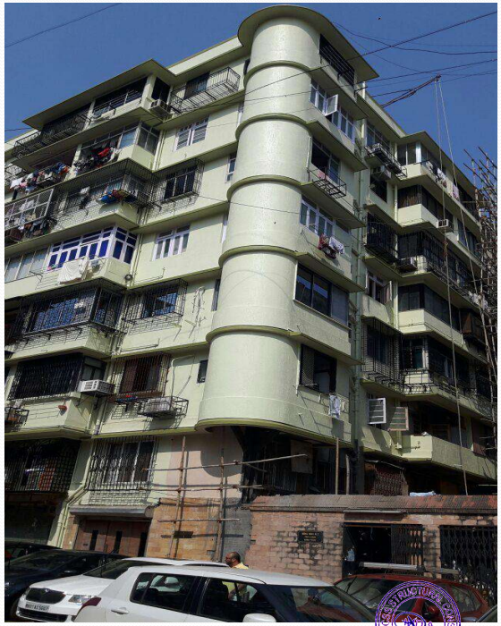 Multi-story residential building with balconies and clothes hanging, parked cars in front under clear blue sky.