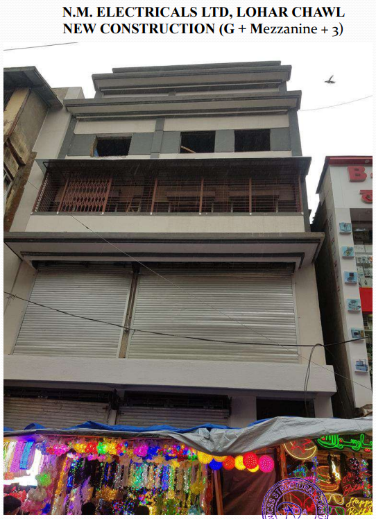 Four-story building under construction with closed roller shutters on lower floors and a colorful market stall with decorative lights in front.